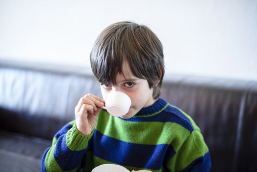 child plays, drinking tea on the sofa at home, lit by the window