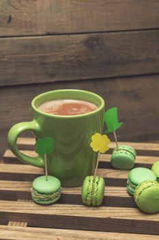 Hot cocoa in green cup and green macaroon cookies scattered on the wooden surface with St. Patrick’s Day attributes. Tonted photo. Shallow depth of field. Copy space