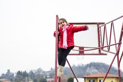 little girl climbs on iron frame of a basketball hoop in an outdoor basketball court