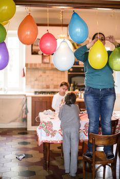 mother with children hang colorful balloons for birthday party in her home, mother standing on the chair hangs balloons at the door