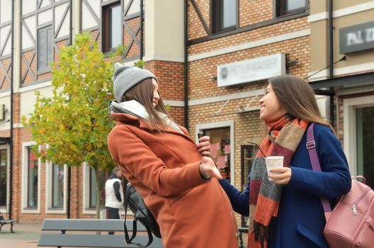 The two girlfriends laugh standing in the middle of the street, with coffee. They are dressed in coats, with backpacks. Side by side to the camera