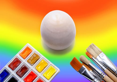 Wooden not painted egg on the background of a rainbow and a brush with paints, the concept of coloring the world with paints.