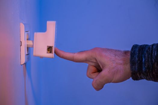 Man press with his finger on WPS button on WiFi repeater which is in electrical socket on the wall. The device help to extend wireless network in home or office.