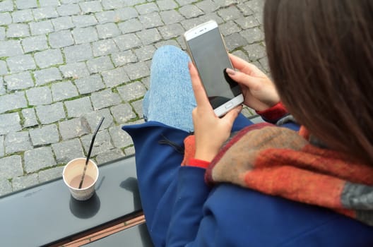 The woman is sitting on the bench and holds the touch phone in her hands. Next is a cup of coffee. The woman is dressed in a coat. Top view