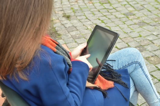 A woman is seated on a bench and uses a black tablet. Side view. Dressed in a coat and jeans. Loose hair
