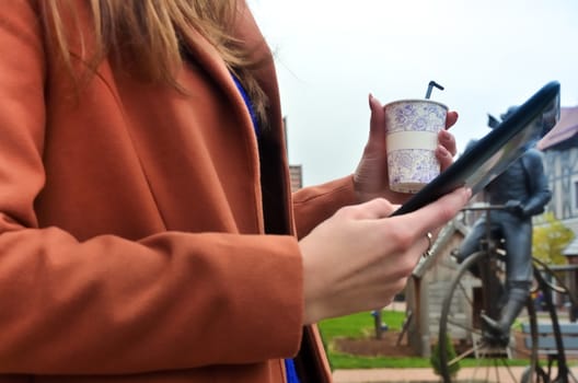 A cup of coffee and a black tablet in the  womens hands. Woman standing in the street. Dressed in a coat