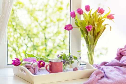 Cozy home concept. Purple fresh tulips in glass vase. Macaroons in glass jar. Cup of hot tea. White tray. Lilac blanket on the windowsill. Sunshine. Coloring and processing photo