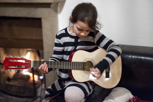 portrait of a 6 year old girl playing guitar at home in front of lit fireplace