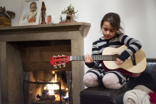portrait of a 6 year old girl playing guitar at home in front of lit fireplace
