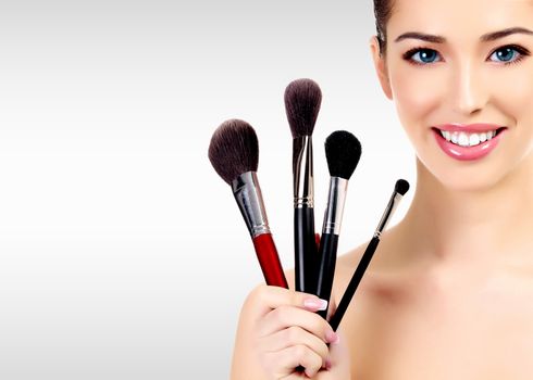 Beauty portrait of lovely beautiful happily smiling woman holding a bunch of make-up brushes against a grey background with copyspace