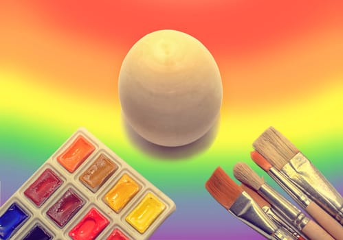 Wooden not painted egg on the background of a rainbow and a brush with paints, the concept of coloring the world with paints.