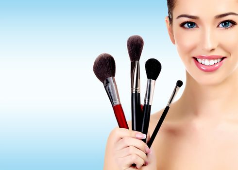 Beauty portrait of lovely beautiful happily smiling woman holding a bunch of make-up brushes against a light blue background with copyspace