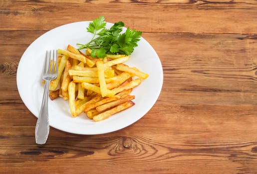 French fries with several twigs of parsley on the white dish and fork of stainless steel on a surface of old wooden planks 
