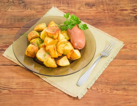 Serving of the country style fried potatoes sprinkled by chopped dill, fried wieners and twig of parsley on dark glass dish and fork on textile napkin on a surface of old wooden planks
