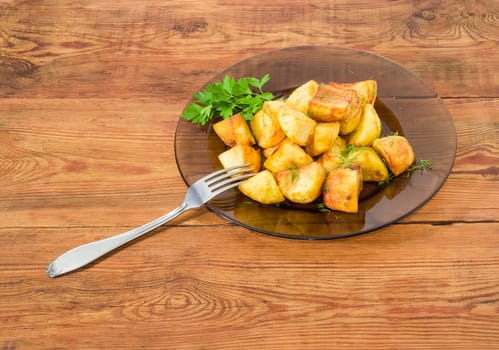 Serving of the country style fried potatoes sprinkled by chopped dill and twig of parsley on dark glass dish and fork on a surface of old wooden planks
