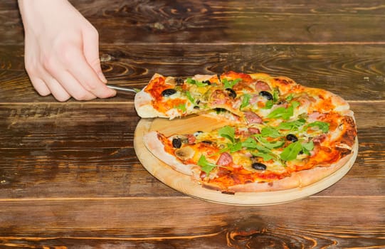 Cooked round pizza with sausages, mushrooms, olives, arugula and slice of pizza on the pizza spatula in the hand of a teenager on the old wooden table
