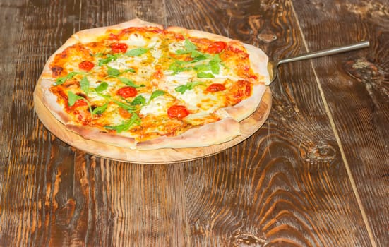 Cooked round pizza with tomatoes, cheese and arugula and pizza spatula on the old wooden table
