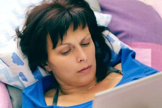 Middle age woman resting in bed with tablet computer and reading news