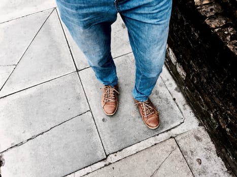Man in blue jeans and brown shoes on the footpath near brick wall.