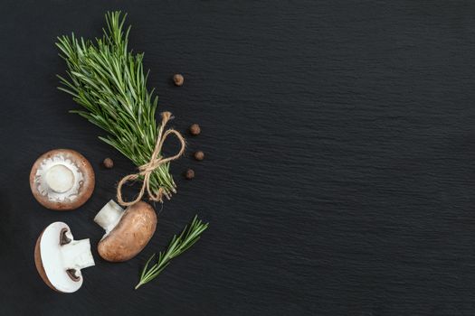 Rosemary bunch of bouquets, mushrooms and fragrant pepper on black stone surface. Top view, copy space.