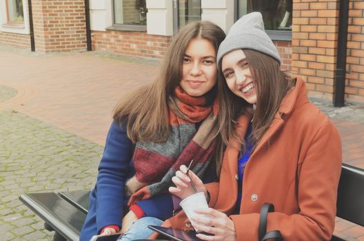 Two young women sitting on the bench outside, smiling at the camera. One of them holds a paper cup of coffee in her hands, and a black tablet. Dressed in coats, brunettes. Europe and Asian