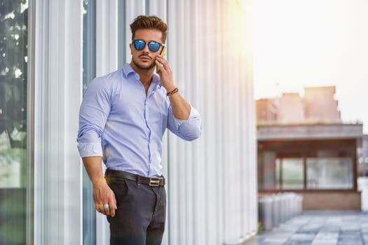 Handsome trendy man wearing white shirt standing and talking on cell phone, outdoor in city setting in day shot
