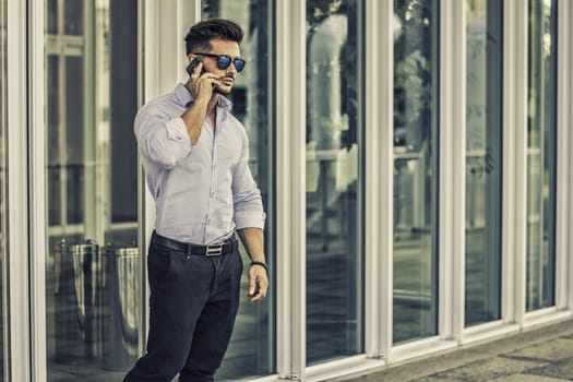 Handsome trendy man wearing white shirt standing and talking on cell phone, outdoor in city setting in day shot