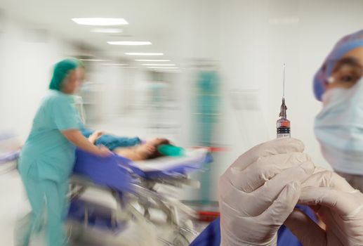 A doctor with syringe and gloves over motion blurred background of nurse in hurry pushing litter down a sterile light hospital corridor.