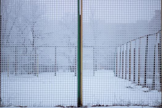 Snowed sport yard behind frosty metal grid net in winter time