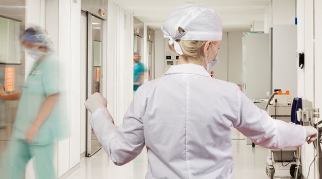 A sterile corridor to operating rooms in modern hospital with female doctor over motion blurred figures of doctors and nurses.