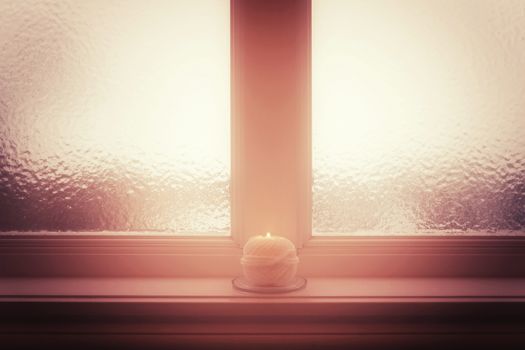 Frosted window and burning candle and in misty light. Winter mood.