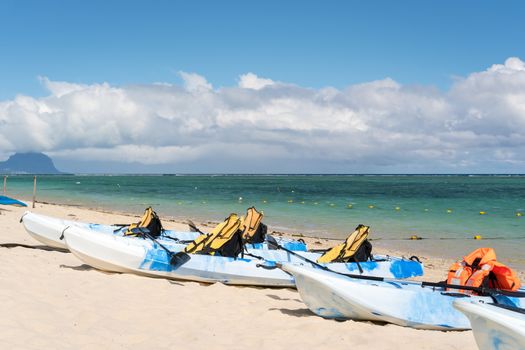 Blue and white kayaks with life jackets on sandy beach at Mauritius island,sunny day with clouds in the sky and mountain background.