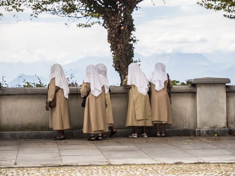 Back view of religious nun community with heads covered standing on stone terrace exploring landscape.