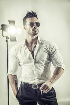 Young macho in formal shirt and sunglasses holding cigarette posing confidently in studio.