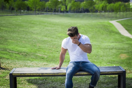 Depressed, sad muscular man in city park in a nice summer day, crying while sitting on bench