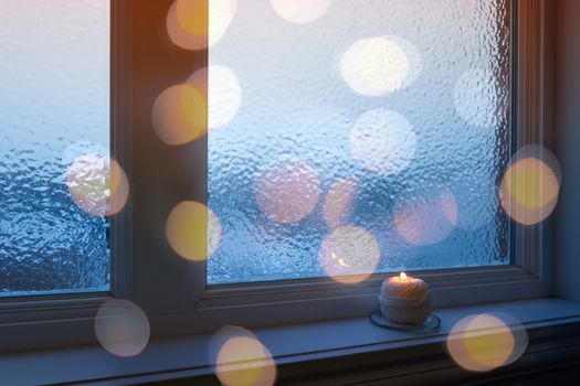Frosted winter window, burning candle and golden bokeh lights.