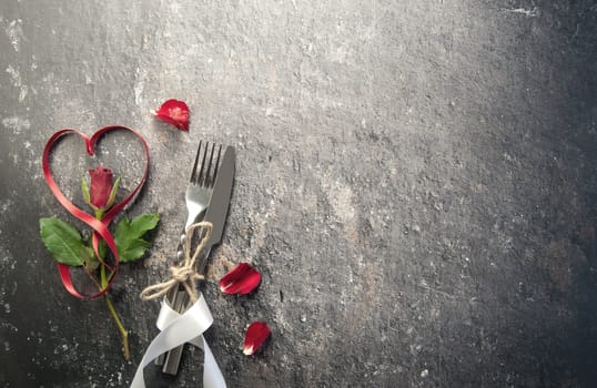 Heart shape satin ribbon with cutlery, rose petals over a concrete marble background