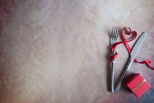 Red ribbon in the shape of a heart inbetween cutlery fork and knife with gift box