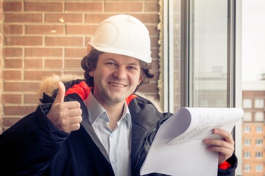 Handyman giving thumbs-up. Engineer with blueprint on brick background. Happy builder with pleased smile. Soft focus, toned