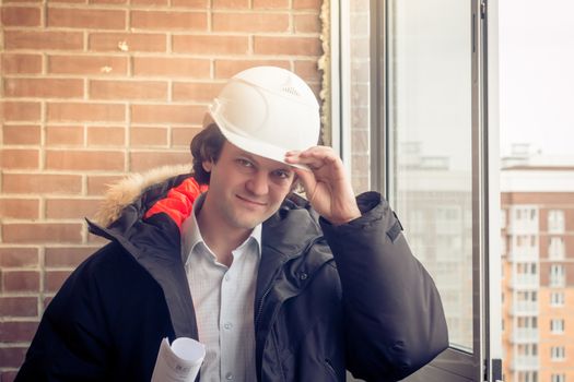 Smiling business engineer with white safety helmet holding blueprint project plan on brick building background, happy businessman greeting gesture and looking to the camera. Soft focus, toned