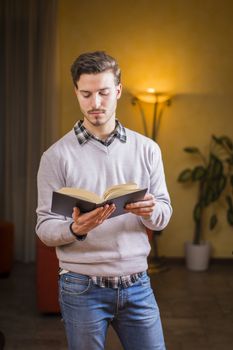 Handsome young man reading book at home in his living-room, standing