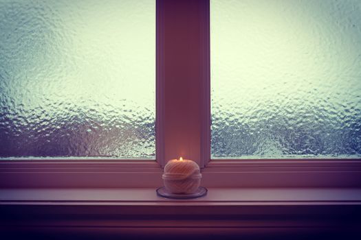 Burning candle and frosted window in twilight. Cozy winter evening.