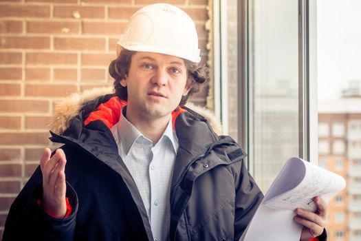 An angry disgruntled builder worker in a helmet with project drawings plans in his one hand and mobile phone in another hand screams on someone. Bad executed work. Aggressive man. Soft focus, toned