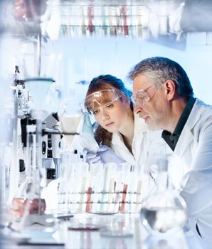Attractive young female scientist and her senior male supervisor looking at the cell colony grown in the petri dish in the life science research laboratory.