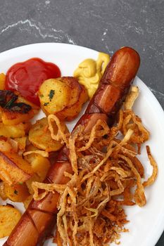 Close up portion of one big grilled sausage with homemade roasted potato, fried onion rings, ketchup and mustard on white plate over grey table, elevated top view, directly above