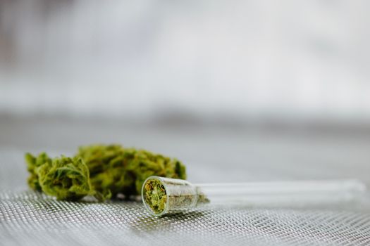 Transparent small glass pipe and dry green marijuana leafs on silver background.