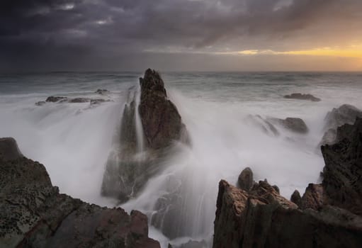 Stormy sunrise with rain and heavy seas crash over volcanic sea stacks on the coast.