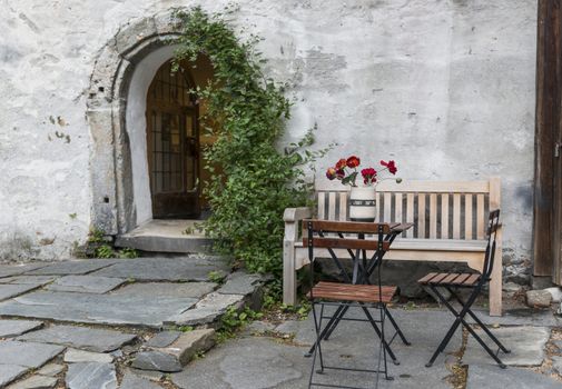 old terrace in bergen norway with red flowers and table and chairs in front of old wall with bow entrance