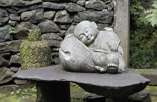 sleeping young buddha statue in garden with green plants and wall with rocks
