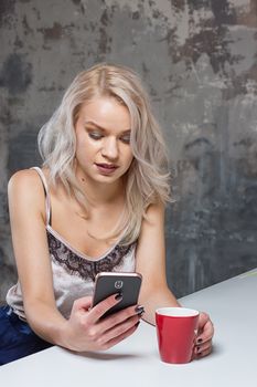 Beautiful blonde girl in home clothes is using a smartphone and smiling while sitting in kitchen